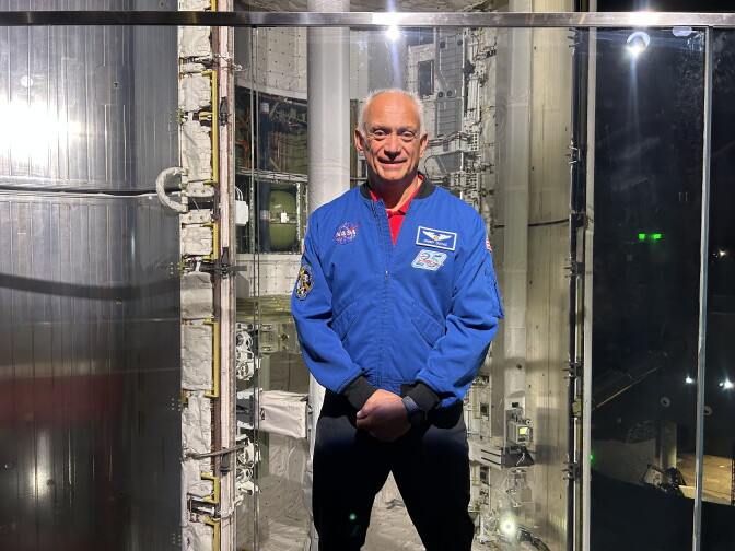 A Latino man wearing a blue long sleeve jacket with the NASA logo on the left breast is standing in front of an opened interior of a space shuttle.