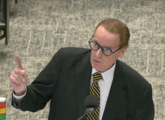 A light skinned man with brown hair wearing a dark suit with a white shirt, yellow and black tie and dark rimmed glasses, points his right finger up while speaking.