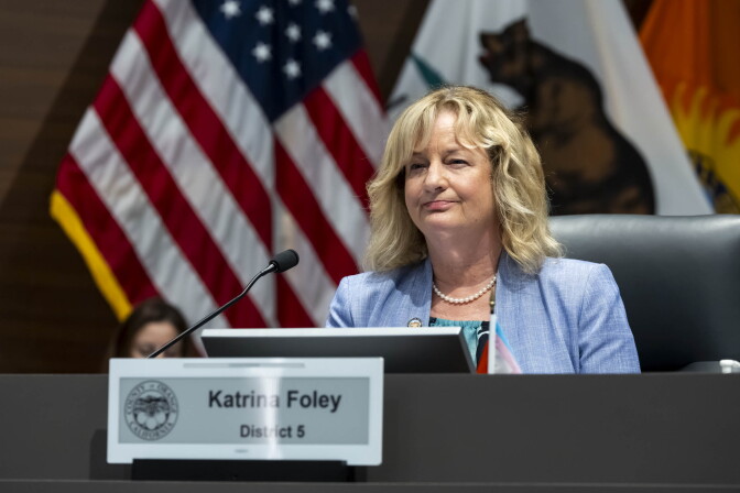 A middle-aged woman with short bond hair and light skin tone wearing a light blue blazer and pearls sits a dais with a microphone pointed towards her. A plaque in front of her reads "Katrina Foley/District 5." She has a slight smile on her face. There's an American flag and a flag of California behind her out of focus.