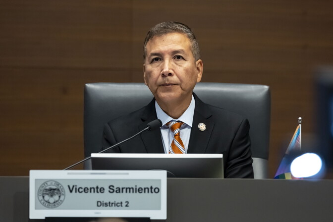 An older man with medium-light skin tone wearing a black suit with a striped orange and white tie sits a dais in front of a plaque that reads "Vicente Sarmiento/District 2."