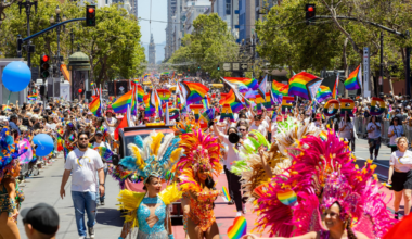 A vibrant parade features people in colorful feathered costumes and crowds waving rainbow Pride flags along a bustling city street.