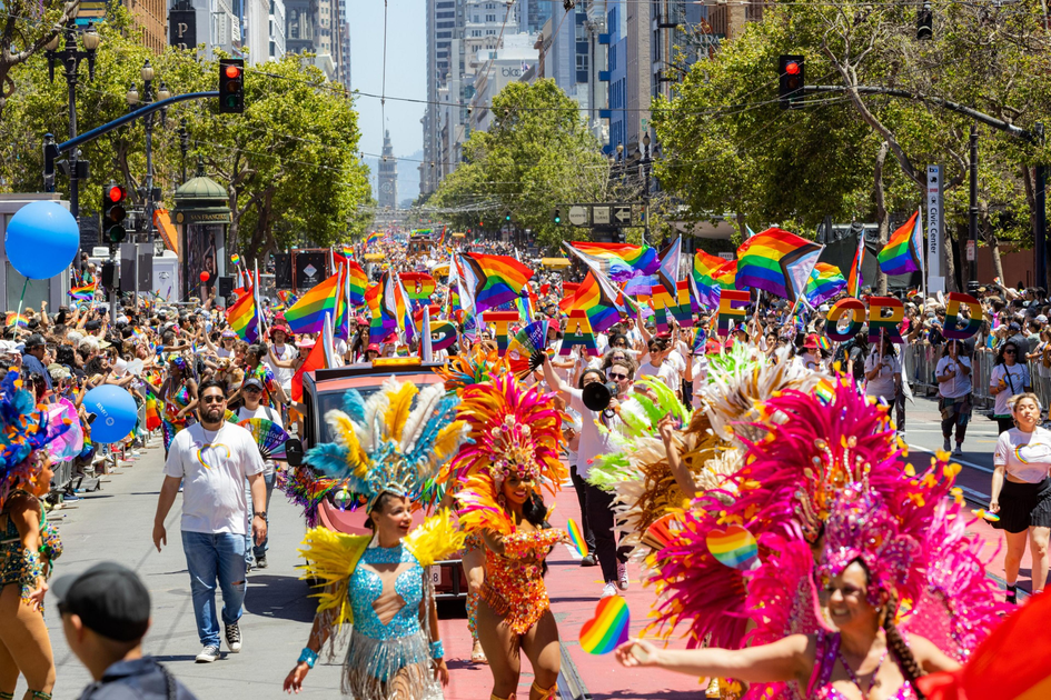 A vibrant parade features people in colorful feathered costumes and crowds waving rainbow Pride flags along a bustling city street.