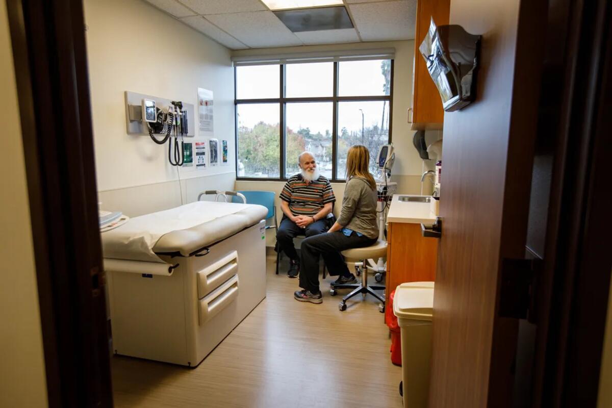 An older man with a beard looks toward a nurse seated next to him. The room includes a medical bed and equipment.