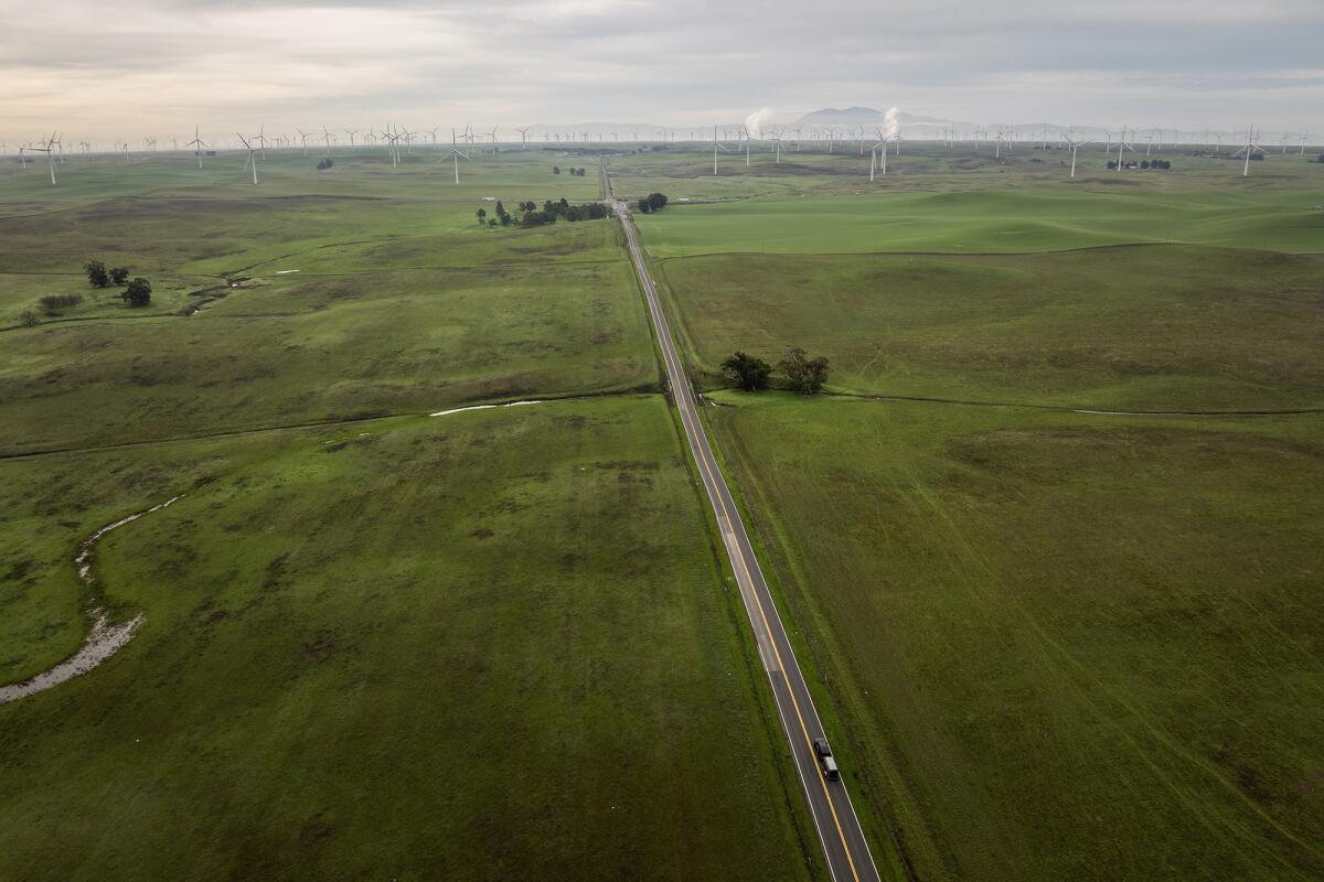 An aerial view shows a two-lane road cutting through expansive green fields with a pickup truck traveling along it.