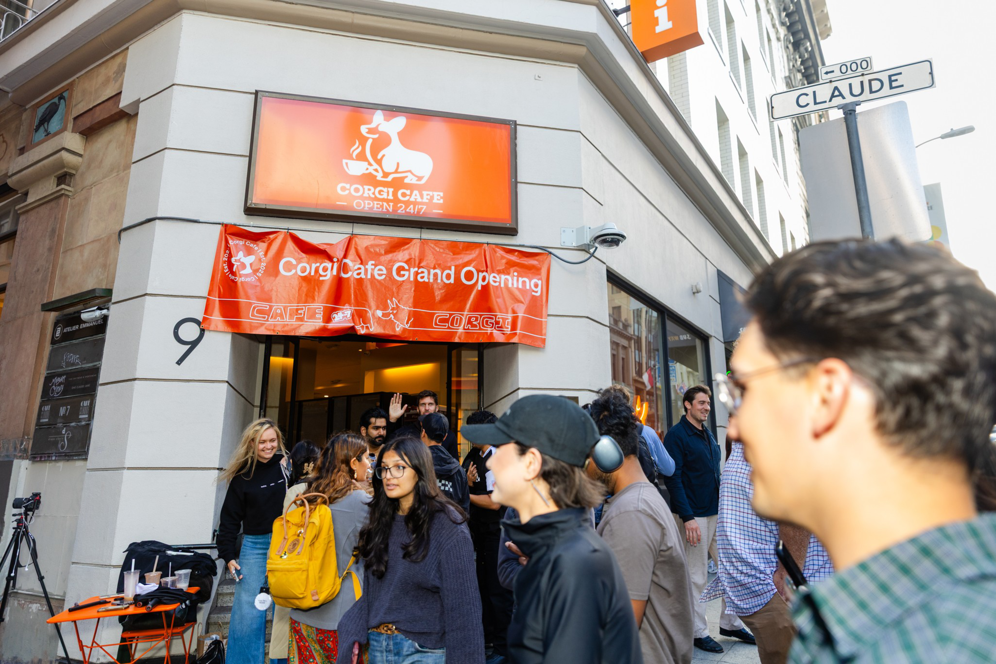 A group of people gathers outside the Corgi Cafe, which has a bright orange sign and banner announcing its grand opening at a corner on Claude Street.