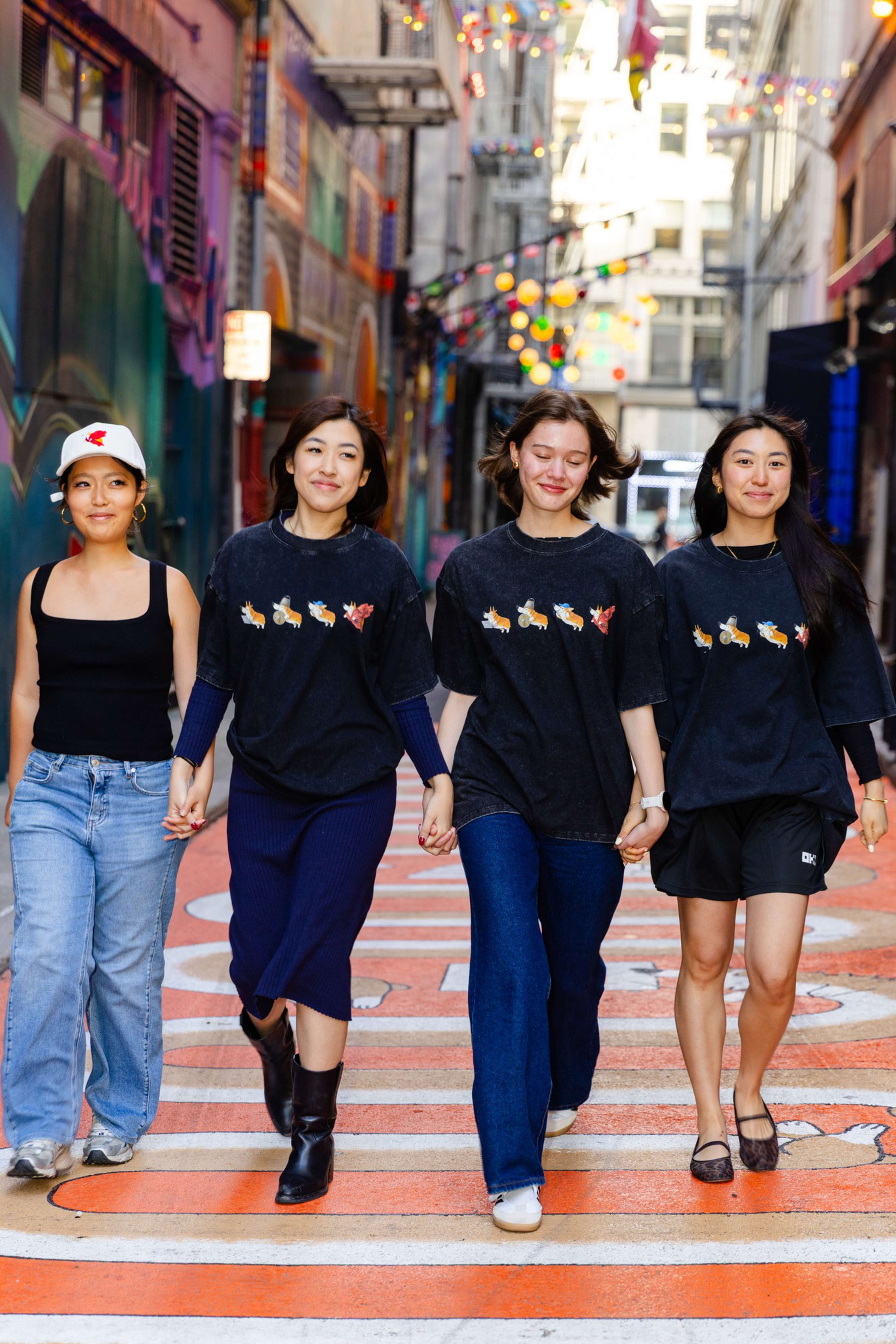 Four young women hold hands and walk down a colorful urban street, three wearing matching black shirts with an animal design, while one wears a black tank top and cap.