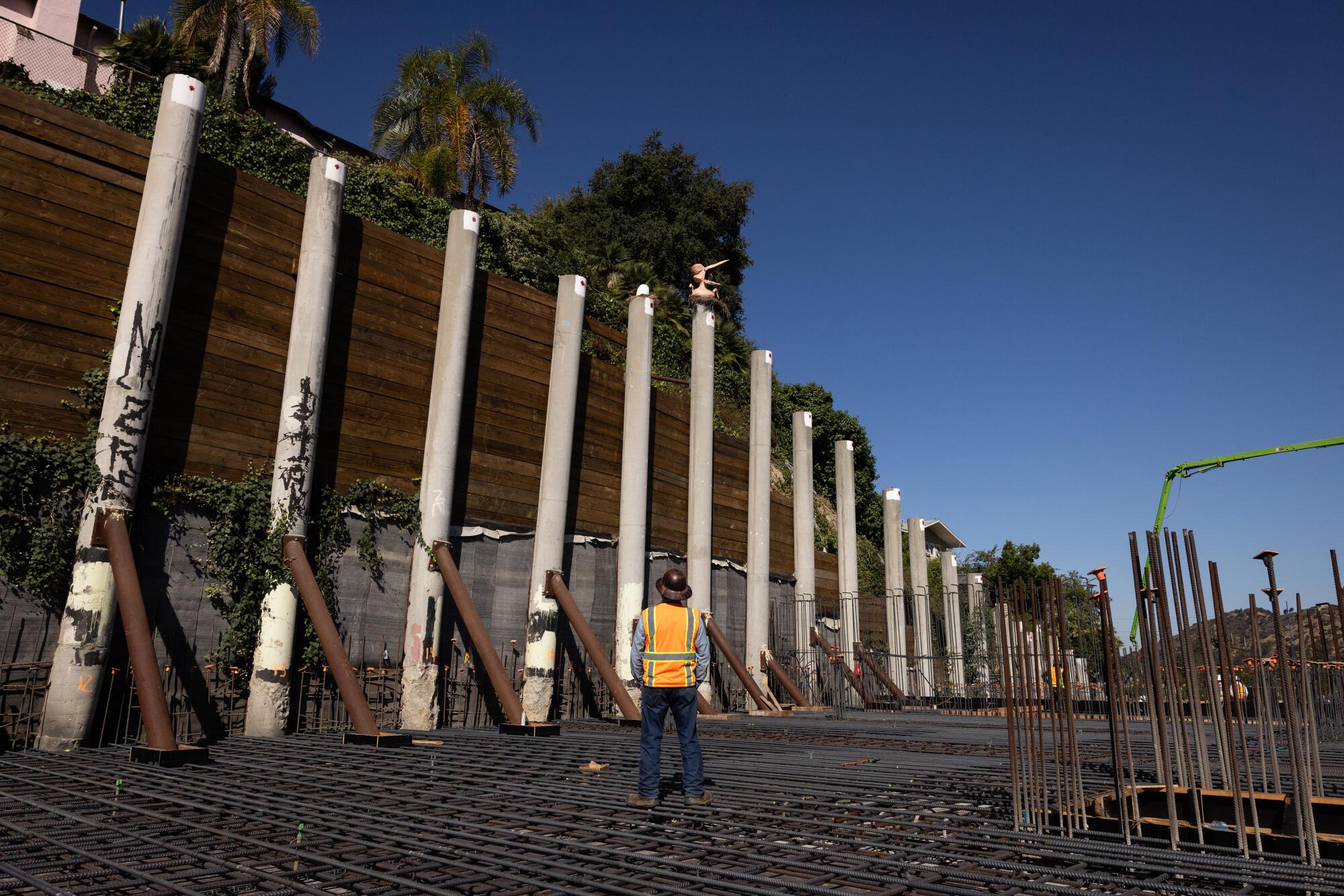 A man in an orange construction vest looks up at a pink bird atop a concrete piller.