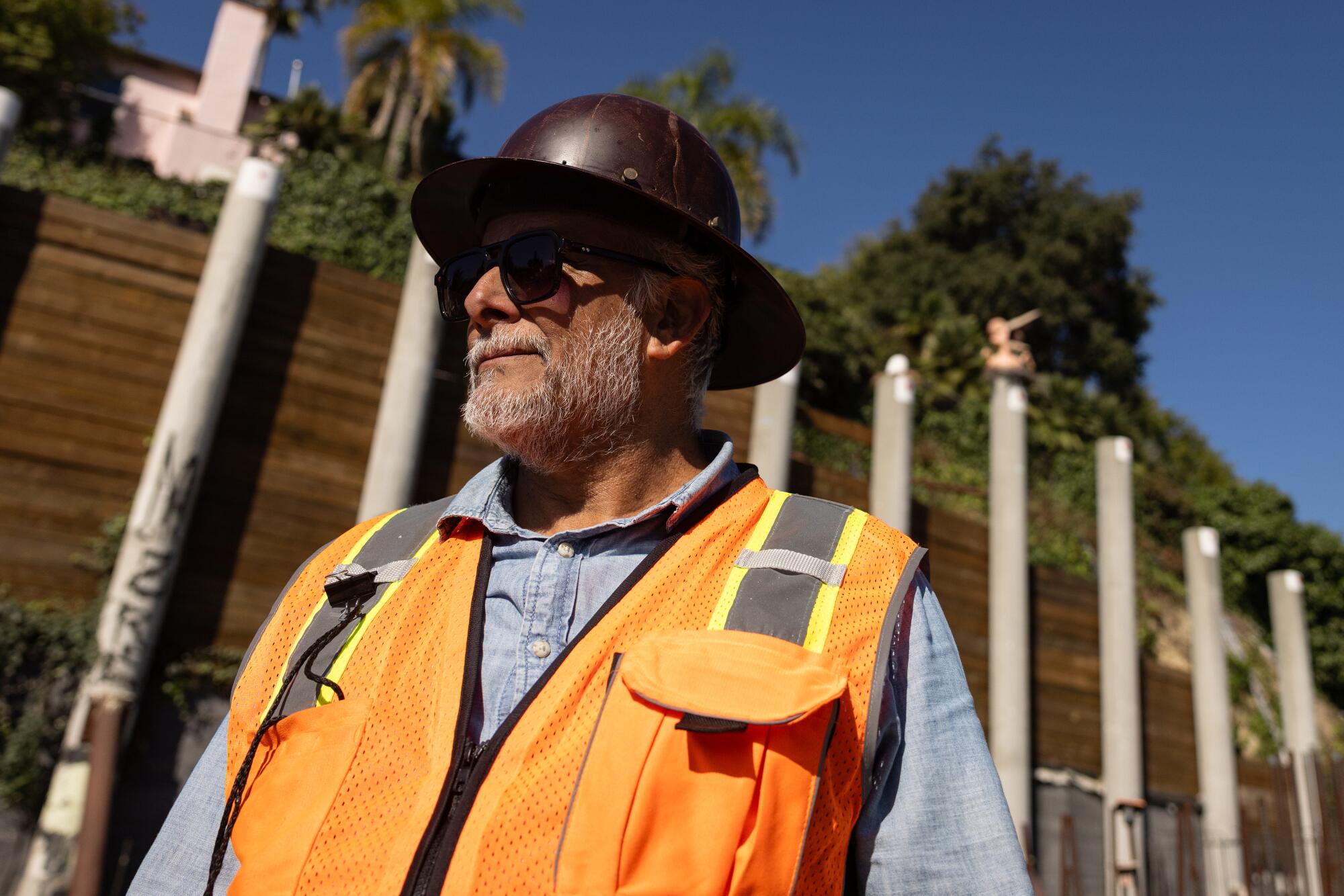 A man in an orange vest poses for a picture as a construction team works in the background.