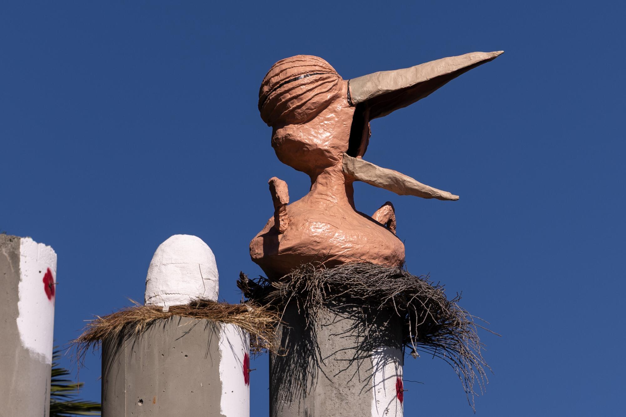 A bird sculpture sits on a nest atop a column with a white egg to its right on another column.