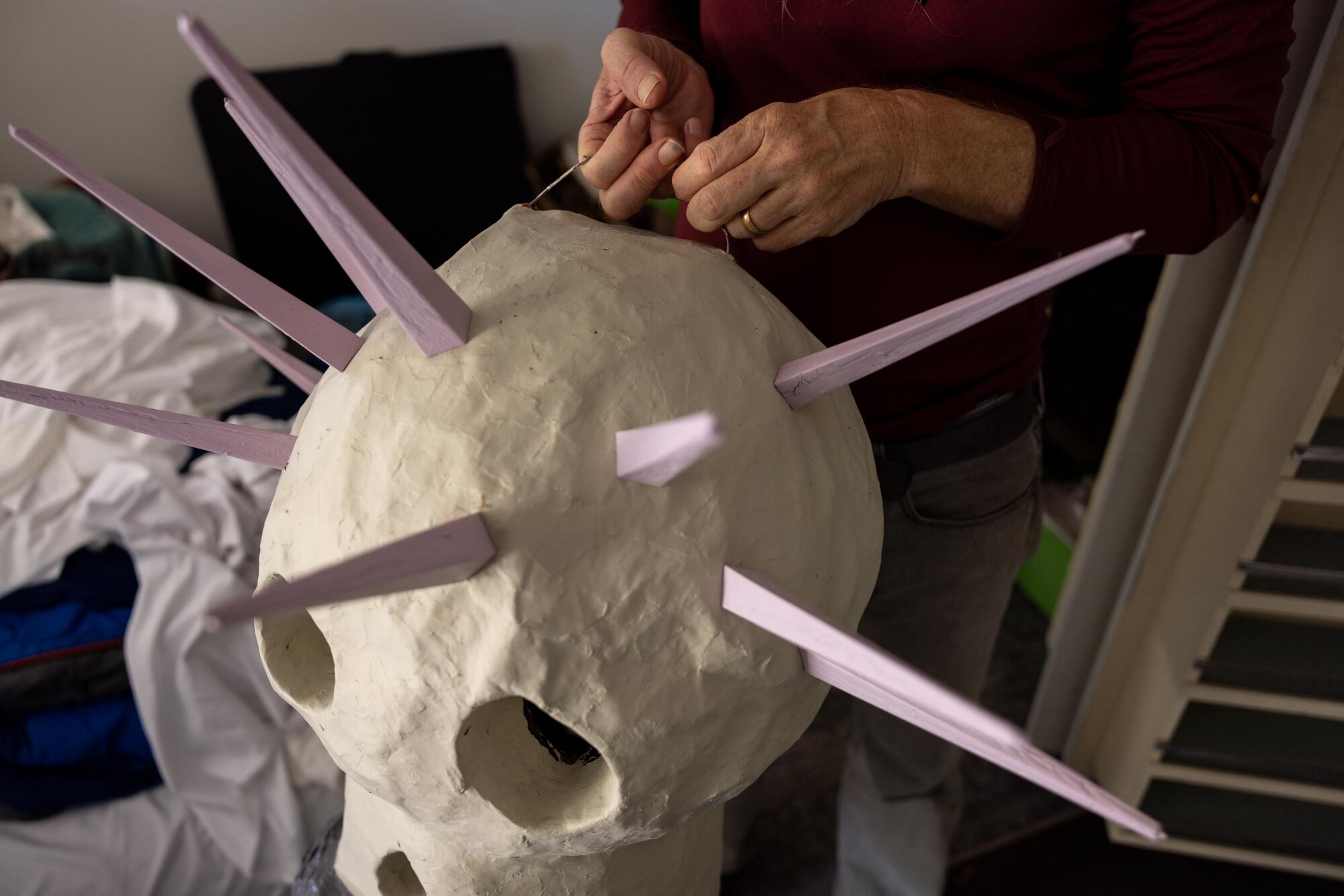 A man's hands hold a string atop a white skull mask adorned with purple spikes.