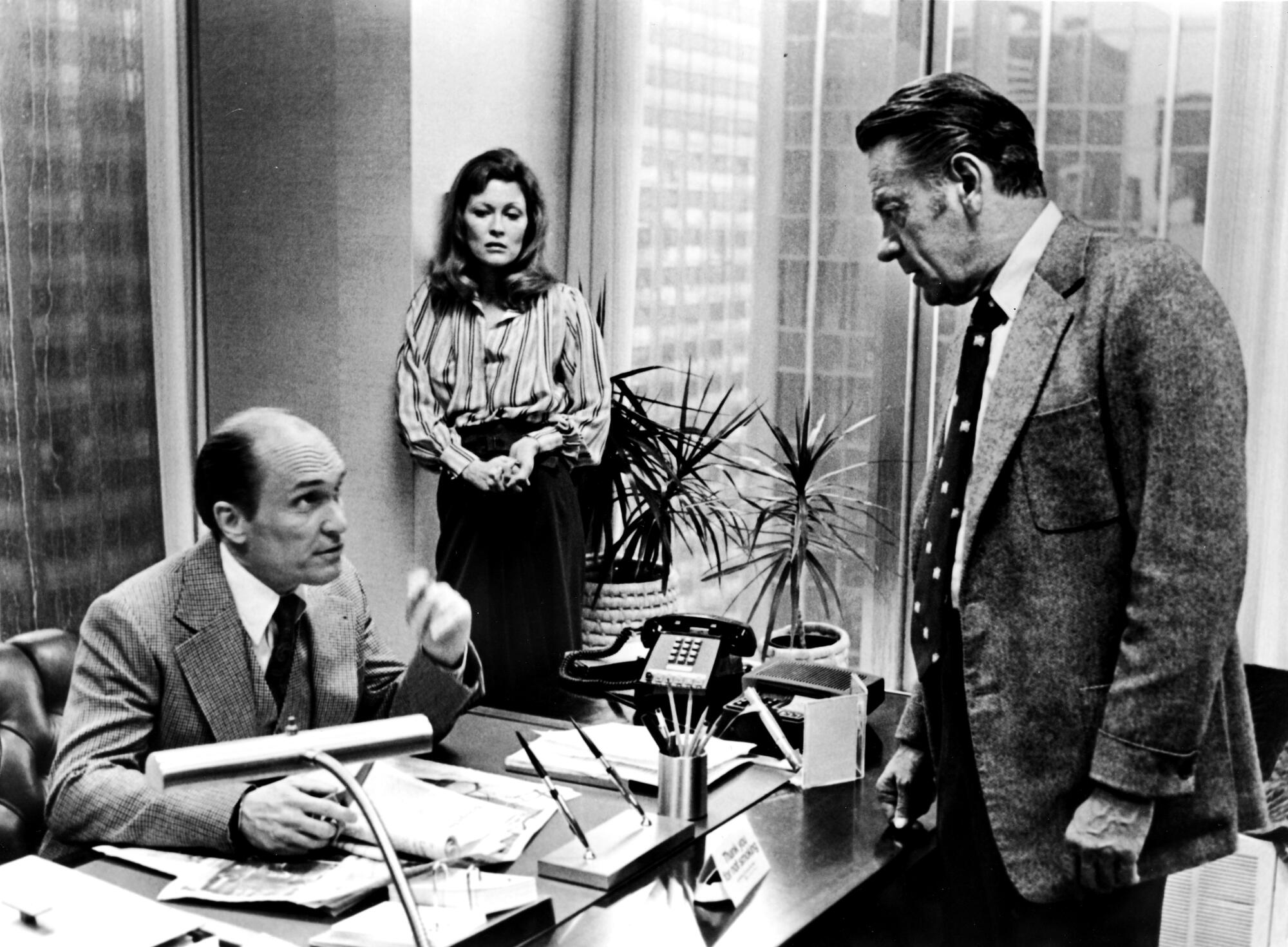 An angry newsman confronts an executive in his office, while a producer looks on.