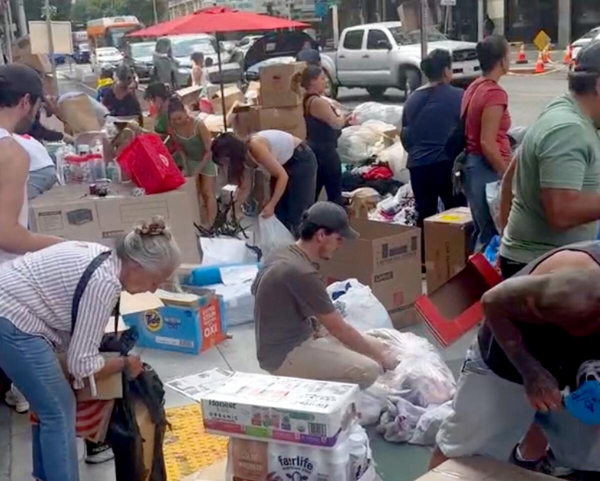 People bend over and kneel alongside boxes and bags of goods on a city sidewalk.
