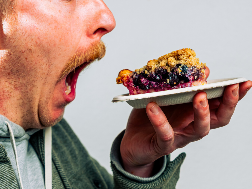 A man with a mustache holds a plate with a slice of berry crumble pie close to his face, looking at it intently.