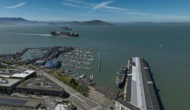 A harbor with docked boats, a large ship near a long pier, a container ship labeled NYK LINE in the bay, and distant hills under a blue sky.