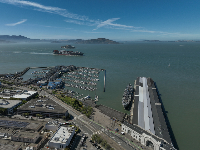 A harbor with docked boats, a large ship near a long pier, a container ship labeled NYK LINE in the bay, and distant hills under a blue sky.