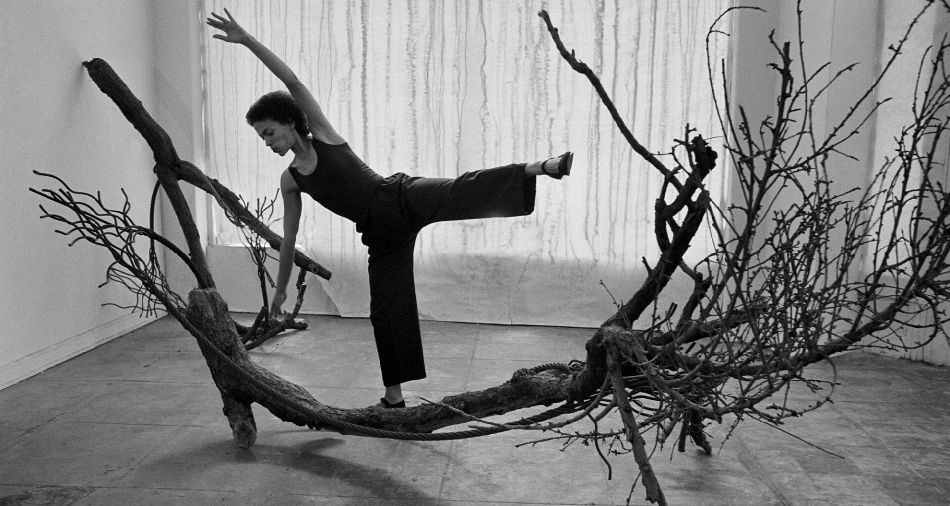 black-and-white image of Black woman posing with leg and arm raised to mimic large tree branch she stands on