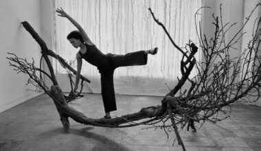 Black-and-white photograph of artist Maren Hassinger striking a balancing dance pose atop a large, gnarled tree branch wrapped in industrial wire rope for her sculpture