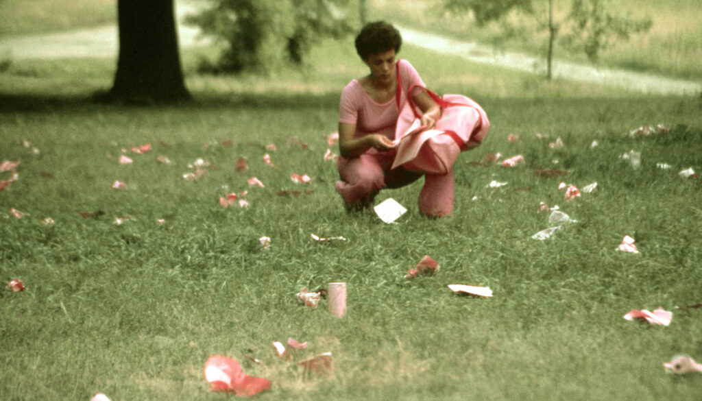 Archival photograph showing artist Maren Hassinger dressed in pink, crouching on a green lawn during a performance. She holds a large pink bag, collecting and scattering pieces of trash that have been painted pink across the grass.