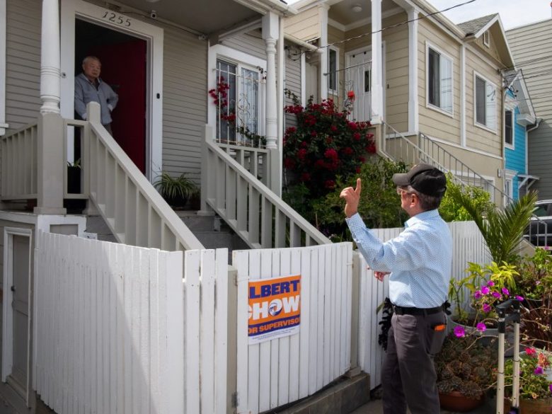 A man stands outside a white fence talking to a person at the door of a house, which has a sign reading "Albert Chow for Supervisor" on the fence.