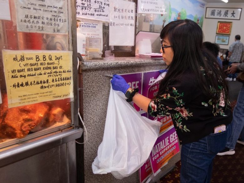 A woman wearing glasses and gloves picks up a plastic bag of food from a counter inside a restaurant, next to a display case of roast meats.