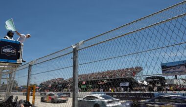 A man in a white hat and shirt waves a green flag while standing in an elevated platform above a tall fence, as race cars accelerate on a track below outdoors during daytime; bleachers with spectators are in the background.