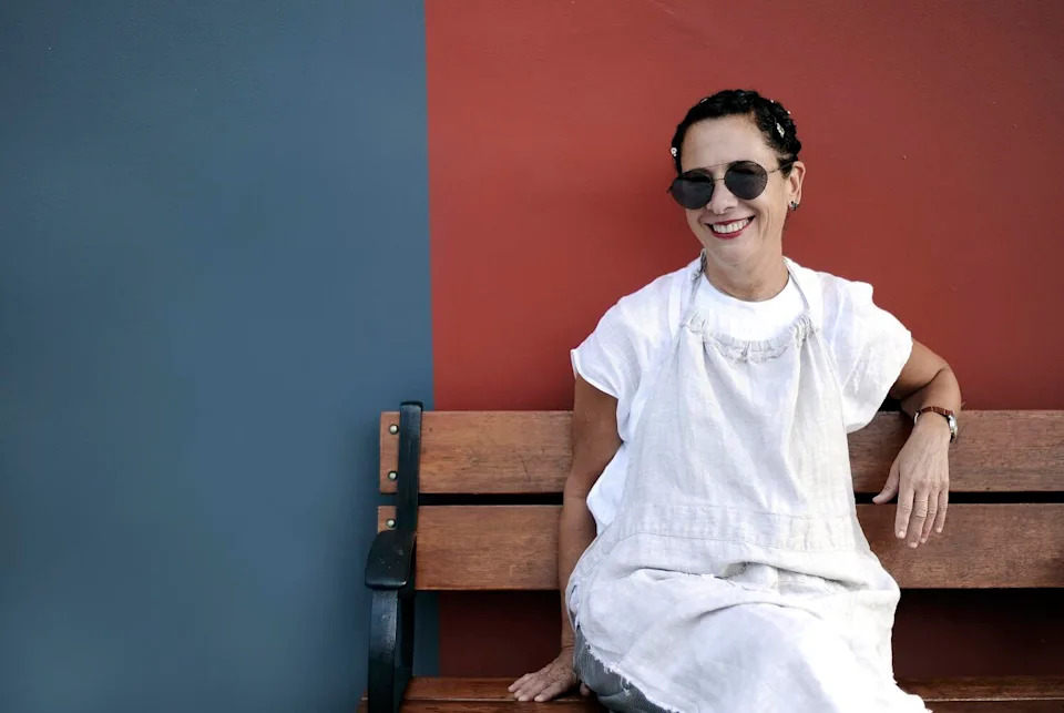 A smiling woman with short, dark hair, in a white dress, seated on a bench