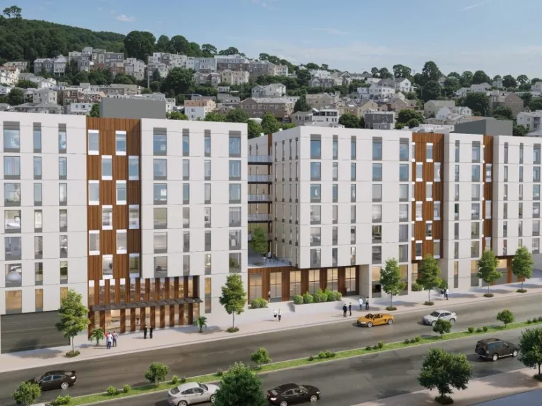 A modern mid-rise apartment complex with white and wood-paneled facades, large windows, and people walking along the sidewalk; a hillside neighborhood is in the background.