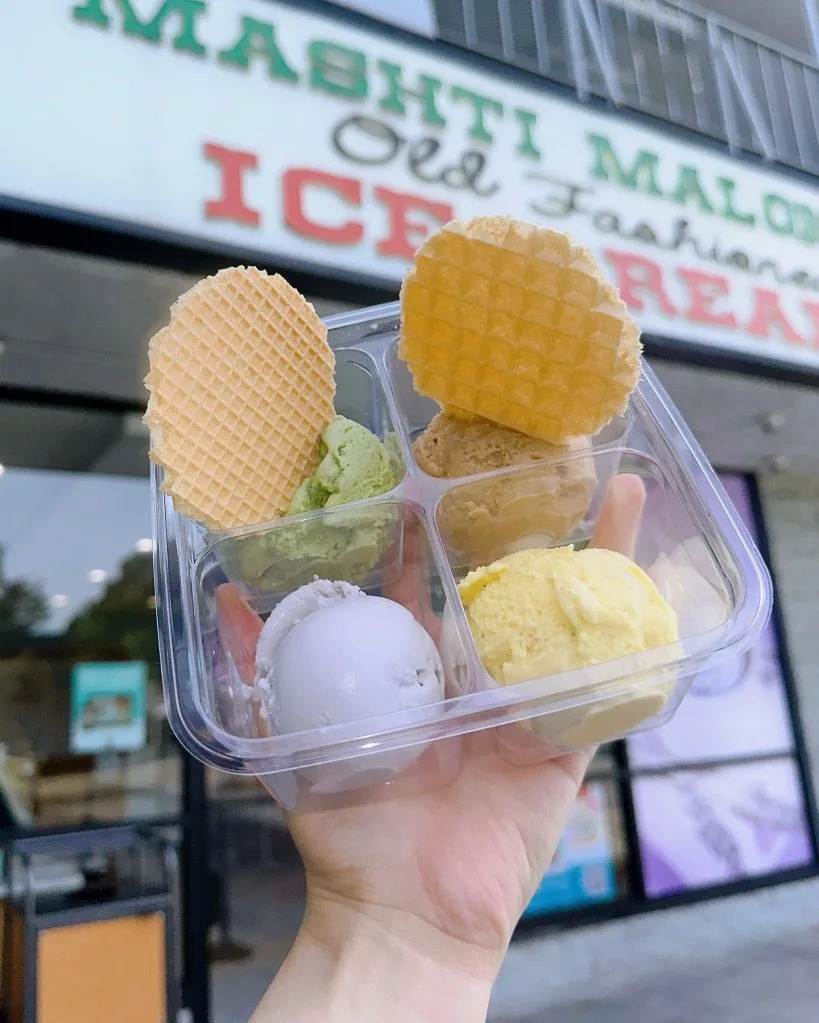 A hand holding a clear plastic container with four scoops of ice cream and two waffle cookies. Instagram/@mashtimalones