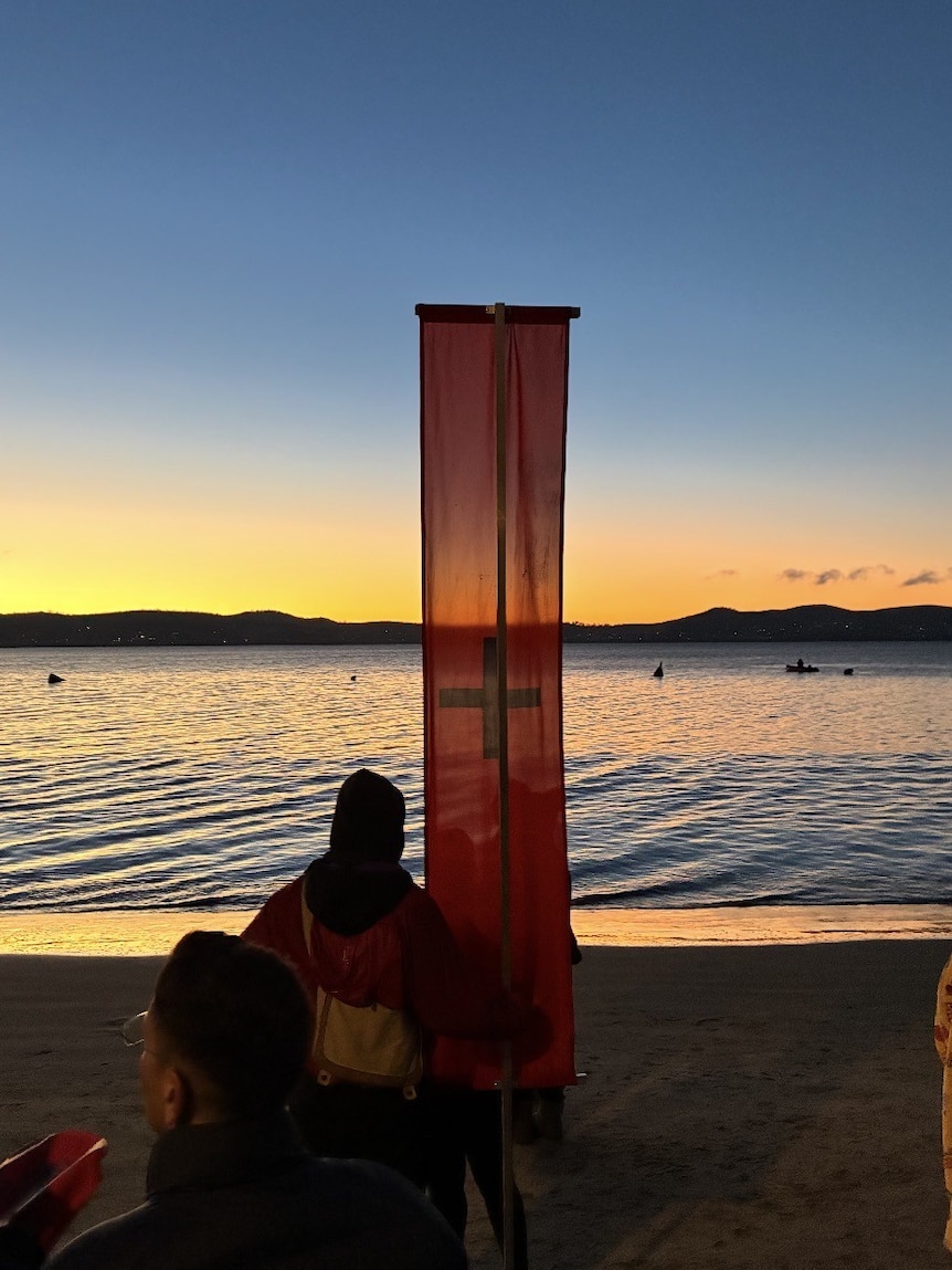 A Dark Mofo red banner with black cross stands on a beach at dawn.
