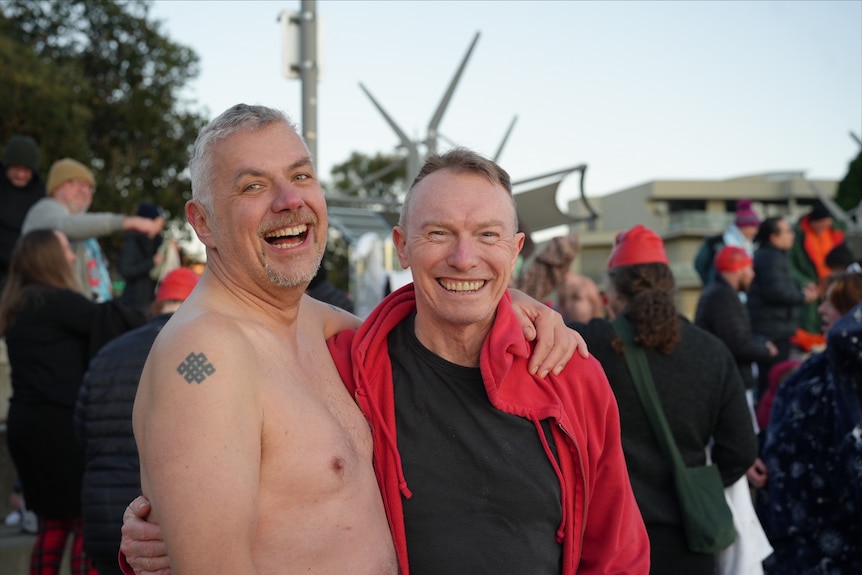 two men stand with arms wrapped around each other, smiling.