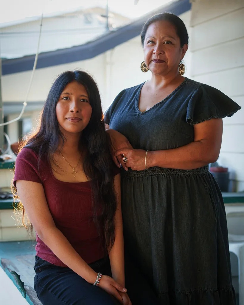 Two people pose together on a porch or patio in warm afternoon light. One person sits on a ledge wearing a dark red shirt and black pants, while the other stands beside them in a dark gray dress with one hand resting on the seated person’s shoulder. Both face the camera with calm expressions.