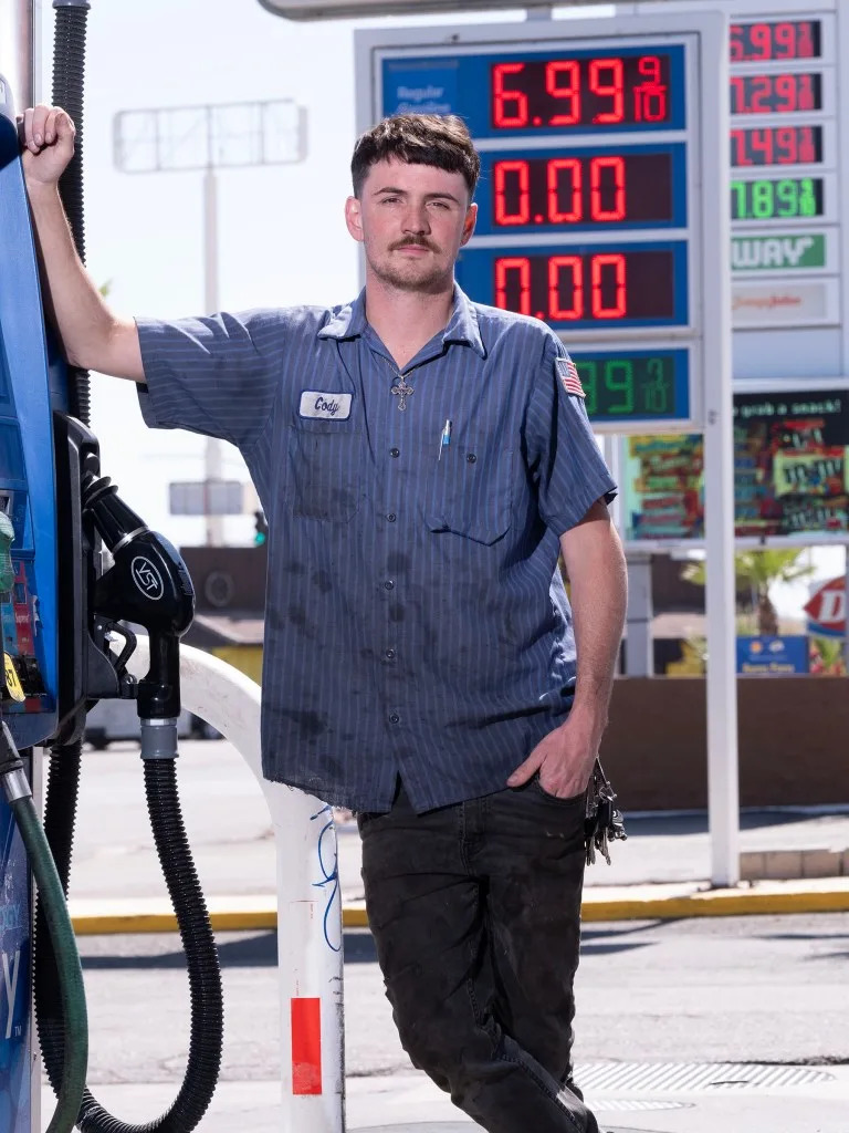 Cody, a Mobil gas station attendant in Needles, CA, stands next to a gas pump and a price sign showing gas at $6.99 per gallon. Andy Johnstone for CA Post
