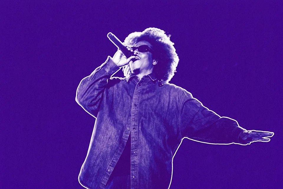 Jay-Z with an afro hairstyle and sunglasses sings passionately into a microphone, wearing a denim shirt, against a purple background. This is a photo from Roots Picnic 2026