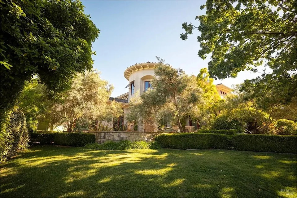 Sunlight filtering through mature trees onto a green lawn in front of a Mediterranean estate