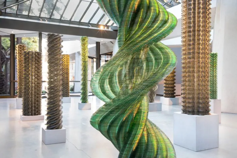 Several tall, spiral sculptures made of stacked glass or translucent material are displayed in a bright, modern gallery with large windows.