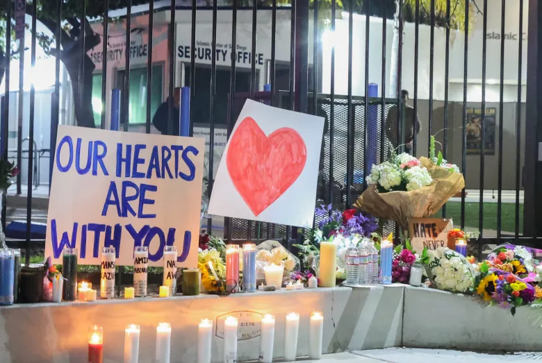 Flowers, candles, and signs outside a mosque. One sign is a large heart, and the other sign says "our hearts are with you."