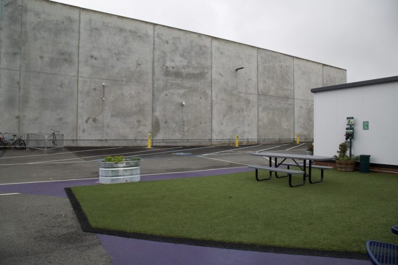 A small artificial grass area with a picnic table is adjacent to a parking lot and a large concrete wall under an overcast sky.