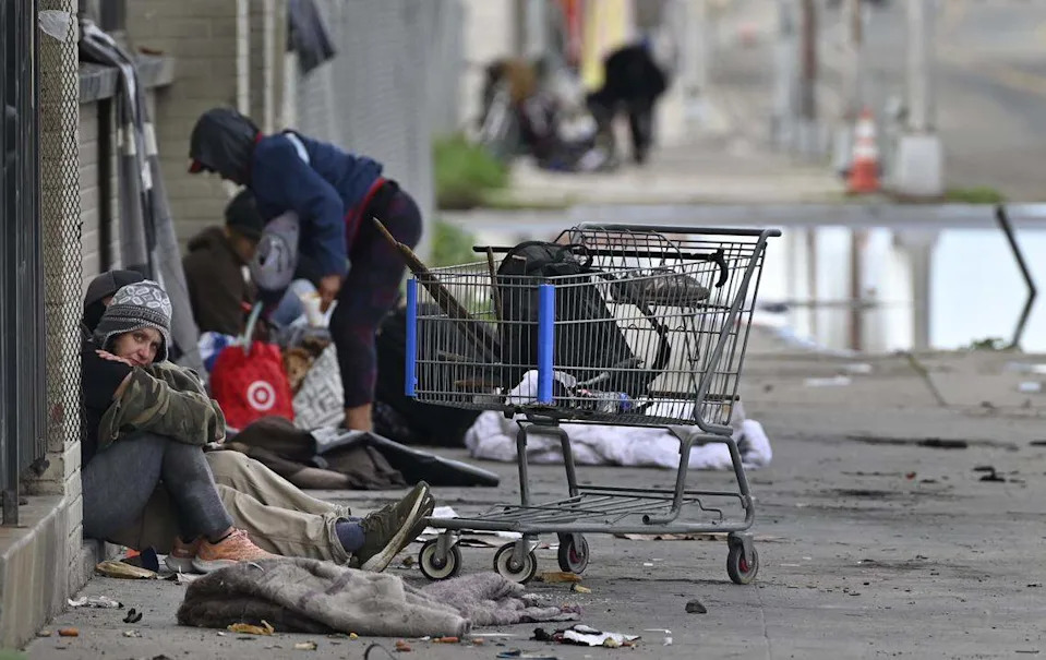 A woman stares as homeless gather on a sidewalk along G Street Friday, Dec. 26, 2025 in downtown Fresno.