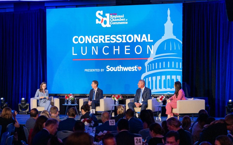 A large screen displays the words "SD Regional Chamber of Commerce Congressional luncheon" with four people sitting in matching airchairs on a stage.