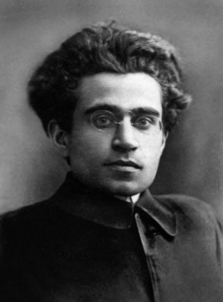 A black-and-white portrait of Antonio Gramsci wearing round glasses and a high-collared jacket with a serious expression.