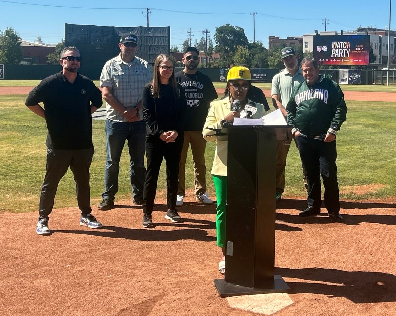 A group of people stand on a baseball field, with one person speaking at a podium while others listen.