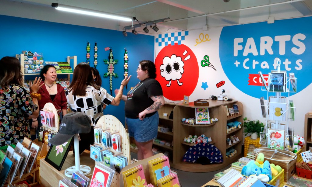 A group of people engage in conversation inside a brightly colored store called "Farts + Crafts," surrounded by whimsical merchandise and playful decor.