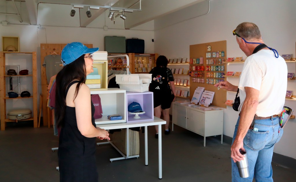 A person in a blue cap and black dress stands at a display table while another person in a white shirt and jeans browses greeting cards on a pegboard wall in a brightly lit retail space.