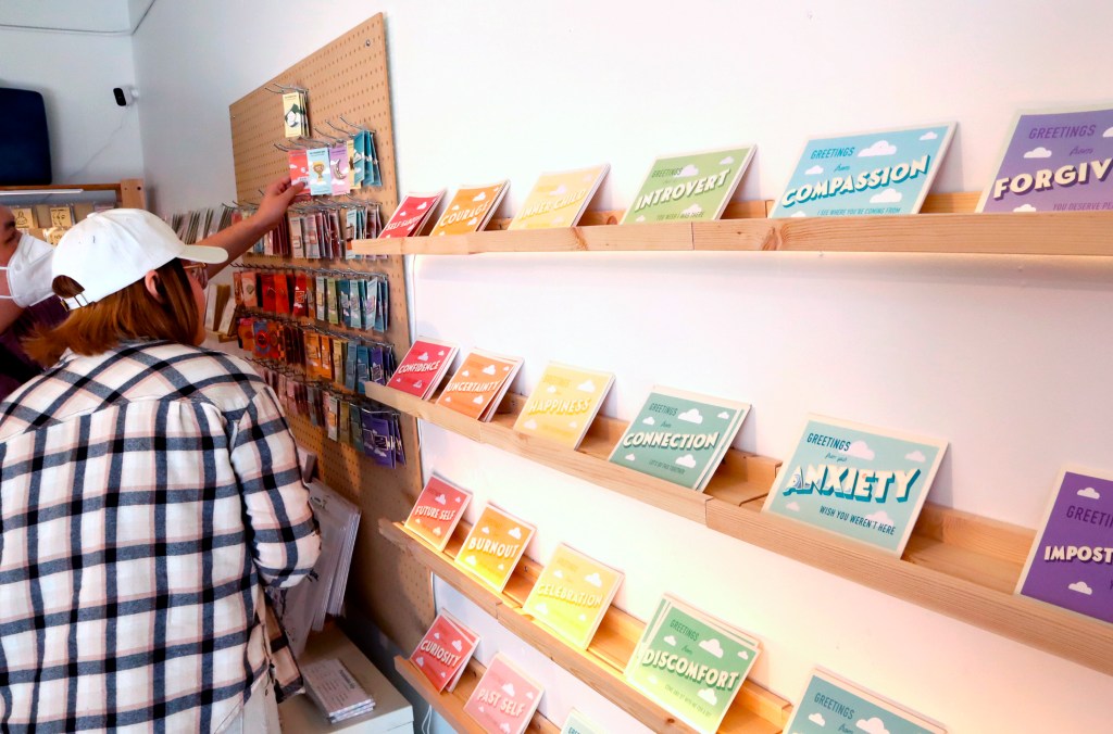 A person in a plaid shirt and white cap browses greeting cards displayed on wooden shelves in a shop.