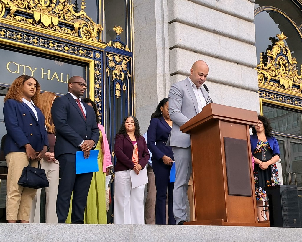 A group of individuals stands on a platform outside City Hall as one person speaks at a podium.