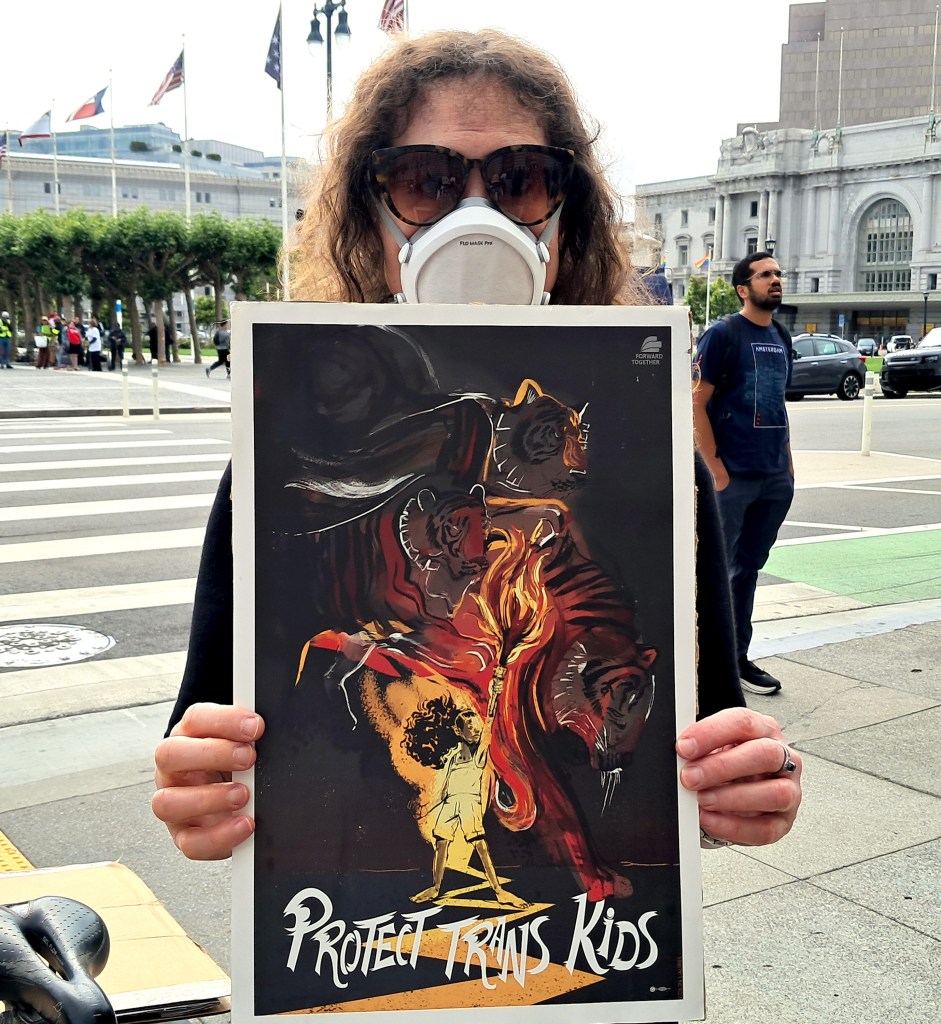 A person wearing a face mask and sunglasses holds a poster that reads "PROTECT TRANS KIDS" in a public square.