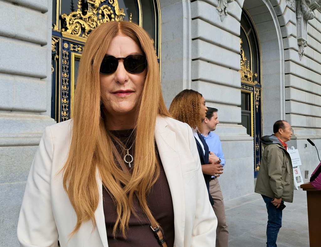 A person with long blonde hair and sunglasses stands in front of an ornate building entrance, wearing a white blazer and a dark top.