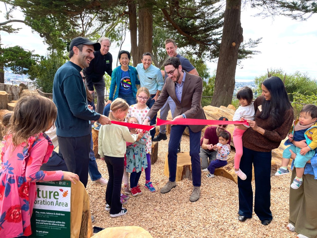 A group of adults and children gather outdoors for a ribbon-cutting ceremony at a nature play area, with some holding the red ribbon and others watching joyfully.