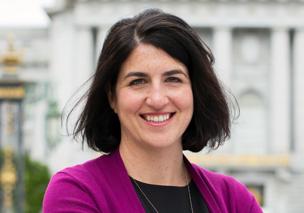 A smiling person with dark hair and freckles wears a vibrant purple cardigan over a black top, standing outdoors in front of a large, light-colored building.