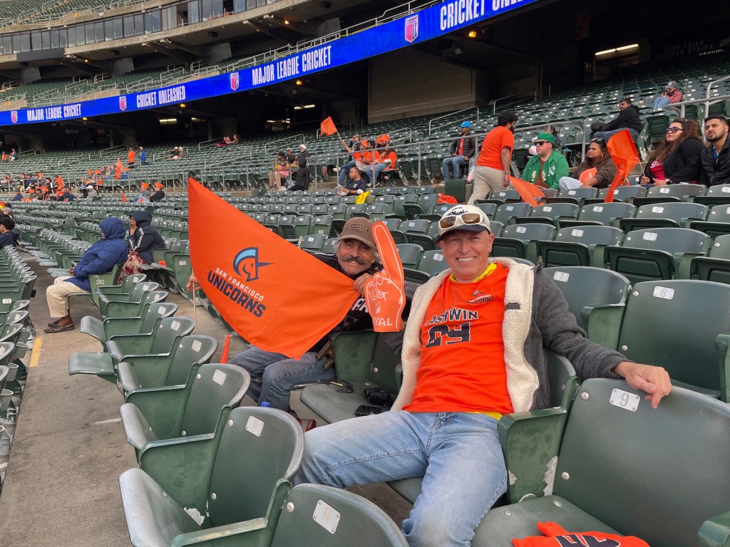 Two fans in orange attire cheer at a baseball stadium, holding a San Francisco Unicorns flag and foam finger.